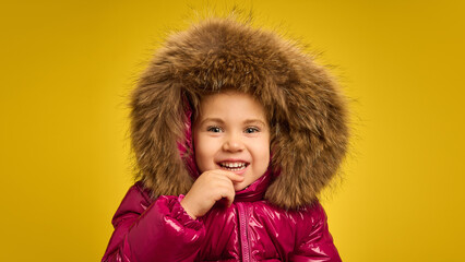 Girl in shiny pink winter coat with fur hood, laughing joyfully, representing happiness and playful fashion, against yellow studio background. Concept of childhood, emotions, lifestyle