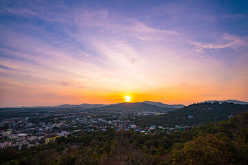 A scenic view of a town nestled among lush green mountains at sunset, with a temple structure perched on the hillside and a vibrant orange sky creating a warm, tranquil atmosphere.