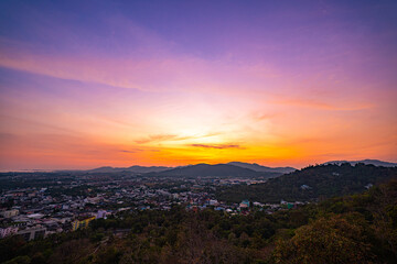A scenic view of a town nestled among lush green mountains at sunset, with a temple structure perched on the hillside and a vibrant orange sky creating a warm, tranquil atmosphere.