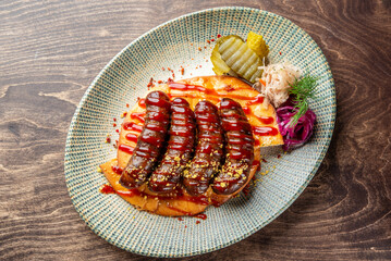 A beautifully plated dish featuring grilled sausages drizzled with sauce, served on a wooden surface. Accompanied by pickles, cabbage, and garnishes for a colorful presentation.