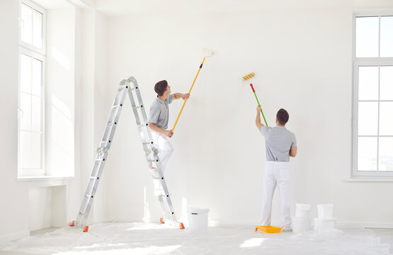 Two male painters in uniform paint a wall white with a roller. Home repair concept. Professional work of builders. Modern apartment renovation, living room interior decoration