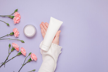 Skincare bottle mockup in woman hand with pink flowers, top view. Mockup facial cream tube, body lotion with copy space