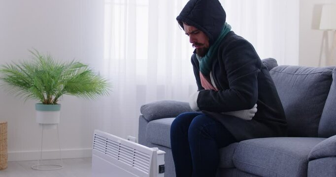 Apartment renter having trouble with heating, cold radiators, dissatisfied man in flat, wearing coat to hide from cold, calling service, apartment management company to complain of low bad temperature
