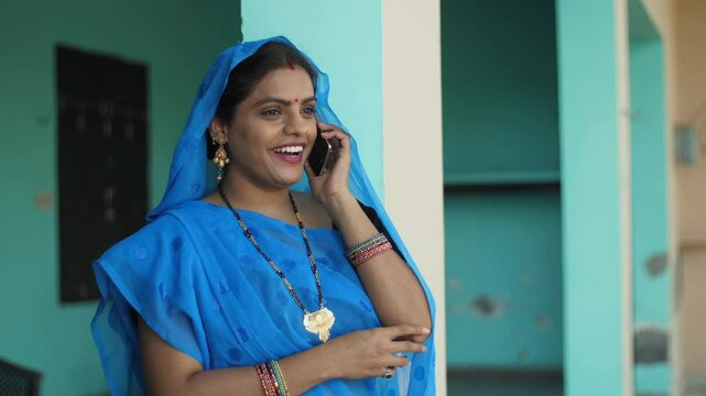 An Indian married woman smiling and talking on the phone at home - independent village woman  modern villagers  rural life. A portrait of a village woman in a traditional saree speaking on the mobi...