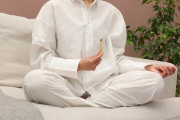 Woman with smoldering palo santo stick meditating on sofa at home, closeup