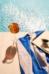 Summer drink with lemon wedge, striped towel, book, and black sunglasses on deck by sunlit pool, evoking peaceful relaxation and warm holiday vibes. Concept of summertime recreation, tourism. Ad