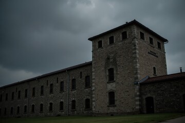 A large, old prison building stands tall with high stone walls and barred windows. In the background, dark clouds gather, creating a moody atmosphere around the structure
