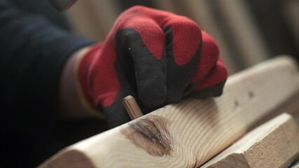 A worker in protective gloves uses a hammer to drive a nail into a wooden plank, showcasing precision and care in woodworking