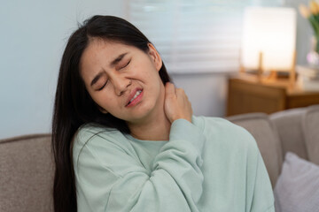 Woman in a mint green sweater grimacing in pain at home, clutching her neck to relieve discomfort and express distress