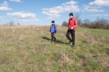 Two young boys walk along a grassy field on a sunny day. The sky is bright blue with scattered clouds. They appear to be enjoying a hike outdoors.