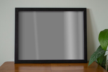 Mockup visualization. Black empty frame standing on wooden table alongside green plant. Poster mock up. Wall art