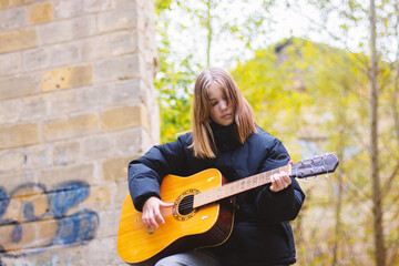 A teenage girl plays the guitar in an autumn park against the backdrop of a destroyed building.
