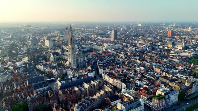 Captured from a high aerial vantage point, the sprawling cityscape of Antwerp is revealed under a clear morning sky, with the iconic spire of the Cathedral of Our Lady prominently piercing the urban