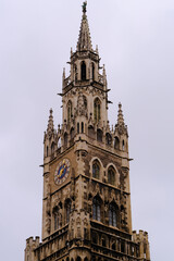 Fototapeta premium A detailed gothic-style tower with a clock, intricate stone carvings, and spires stands tall against a cloudy grey sky, exuding historical elegance and architectural grandeur.