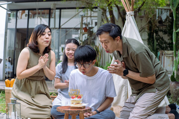 An Asian family celebrating a birthday outdoors. A boy is blowing out candles on a small cake while surrounded by smiling family members, cheerful atmosphere with a natural backdrop