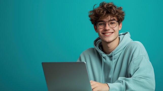 Smiling Young Man in Hoodie Working on Laptop with Bright Turquoise Background