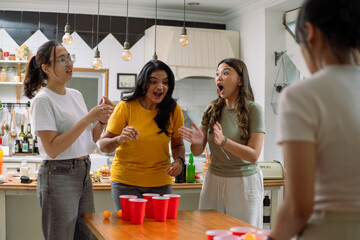 Asian friends enthusiastically playing beer pong in a lively indoor setting. Red cups and orange...