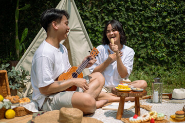 Asian couple enjoying a lively picnic in a serene garden setting. The man plays a ukulele while the...