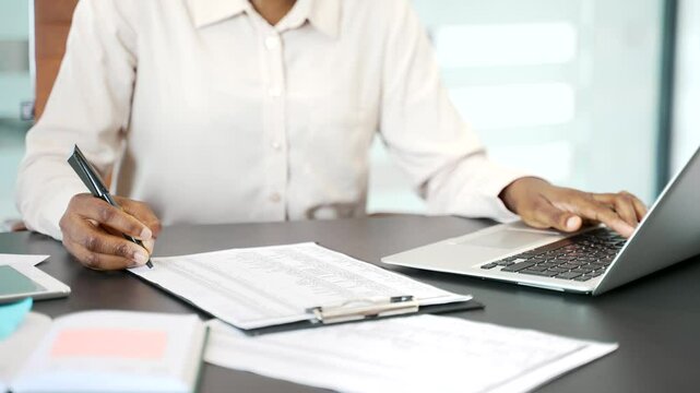 Close up of a female hand filling out documents with a pen using laptop at desk at workplace in business office. African American businesswoman is engaged in paper work, making financial calculations