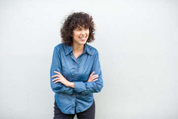 Smiling young woman with curly hair and blue shirt standing with arms crossed against white wall