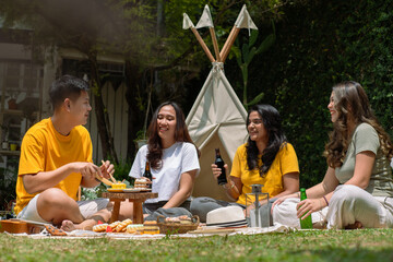 A diverse group of Asian friends enjoying a cheerful outdoor picnic with drinks and snacks. They sit on a blanket near a tipi, sharing laughs amidst a lush green garden under bright sunlight