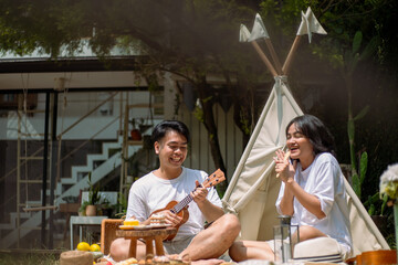 Asian couple enjoying a lively picnic in a serene garden setting. The man plays a ukulele while the...
