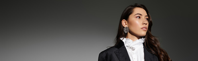 A young woman with shiny brown hair poses confidently in stylish apparel against a grey backdrop,...