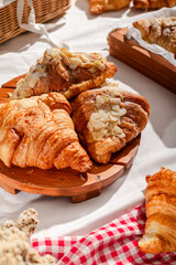 Golden flaky croissants with almond toppings displayed on a rustic wooden board, set on a white tablecloth with a red checkered napkin, creating a cozy and inviting bakery atmosphere