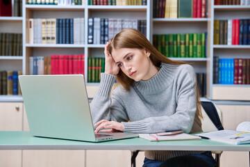 Sad teenage student girl preparing in classroom library looking at laptop
