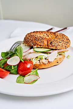 A savory sandwich features sliced chicken and cucumbers nestled in a sesame bagel. Accompanied by cherry tomatoes and mixed greens, it offers a refreshing meal option