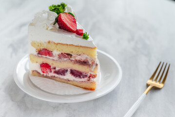 Slice of strawberry shortcake with layers of cream and fresh strawberries, garnished with whipped cream and parsley, served on a white plate with a gold fork on a marble table