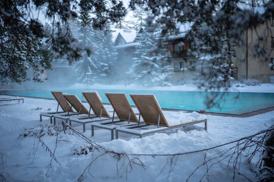 Warm open-air swimming pool with blue water and snow covered loungers in cold weather, outdoor. Winter wellness spa resort. 