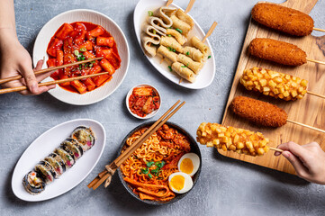 A top-down view of various Korean street foods, including crispy Korean corn dogs, odeng skewers, tteokbokki, spicy ramyeon, gimbap, and kimchi, being enjoyed with chopsticks on a gray surface