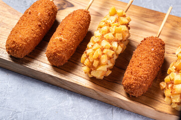 Close-up of crispy Korean corn dogs on a wooden serving board, with variations including traditional breadcrumb coating and potato-crusted versions, placed on a textured gray surface