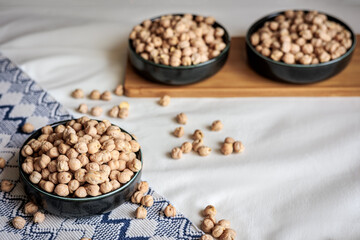 Dried chickpeas in a ceramic bowl on a textured fabric with scattered chickpeas and wooden tray in the background, highlighting natural food and rustic decor