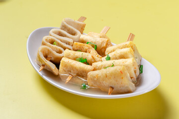 A person holding a white plate filled with Korean fish cake skewers (Eomuk) garnished with chopped green onions, served in a light broth against a vibrant yellow background