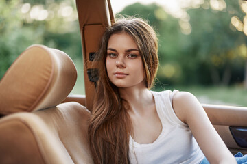 Young woman with long hair in a white tank top sitting in a convertible car, showcasing a serene...