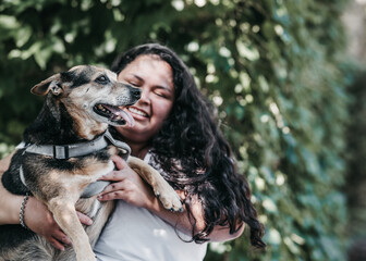 Latina girl holds a medium-sized dog in a gray harness. She has long dark hair and wears a white t-shirt. Background of green foliage, outdoors