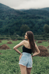 A woman stands against the backdrop of a green field with several haystacks. Wooded hills or mountains are visible in the background and the sky is cloudy.