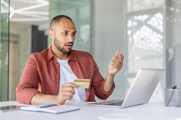 Upset and deceived man at workplace holding bank credit card in hands, received error of funds...