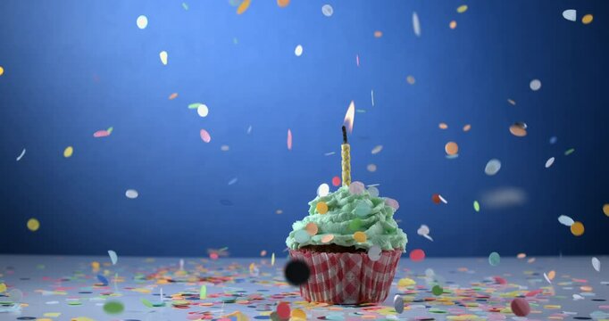 Falling confetti on top of cupcake with one candle on top. Birthday anniversary festive celebration concept on table with blue backdrop