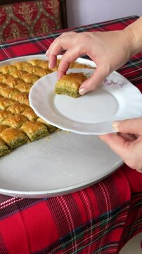 A hand placing a piece of golden, flaky baklava on a plate from a large serving tray, set on a red plaid tablecloth.