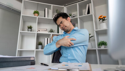 Young Businessman in Modern Office Experiencing Aches and Pains While Working at Desk with Documents and Laptop