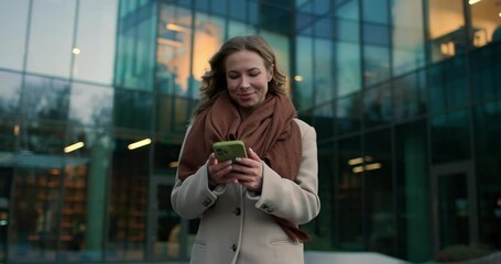 Beautiful Caucasian woman walking away from modern glass building, holding green smartphone and browsing attentively. Typing reply to text. Evening reflections creating dynamic urban atmosphere.