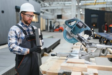 A carpenter cuts a wooden plank on a sawmill in a modular building factory, providing precise cuts for a prefabricated structure