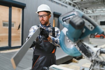 A carpenter cuts a wooden plank on a sawmill in a modular building factory, providing precise cuts for a prefabricated structure