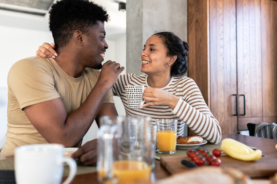 A cheerful couple enjoying a breakfast together at home, smiling and engaging in a playful conversation while seated at a wooden dining table with fresh food and beverages