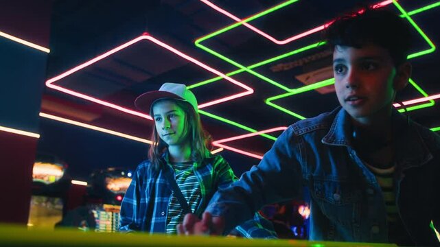 Medium shot of two focused young multiethnic 1980s boys playing game of air hockey at entertainment arcade on weekend, scoring goal against opponents, cheering, jumping and giving high fives