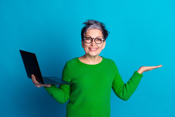 Cheerful senior woman with laptop posing confidently against a bright blue background in casual...