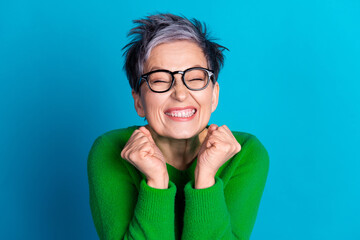 Elderly woman with grey hair and glasses smiling joyfully in green sweater against blue background